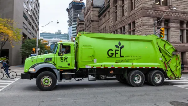 A bright green GFL (Green For Life) garbage truck is driving down a city street, passing a crosswalk. The truck has the GFL logo with a leaf design and website URL “www.gflenv.com” on the side. Office buildings and pedestrians are visible in the background.