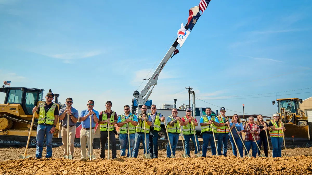 A group of people wearing high-visibility gear stand in front of shovels in the dirt on a bright day.