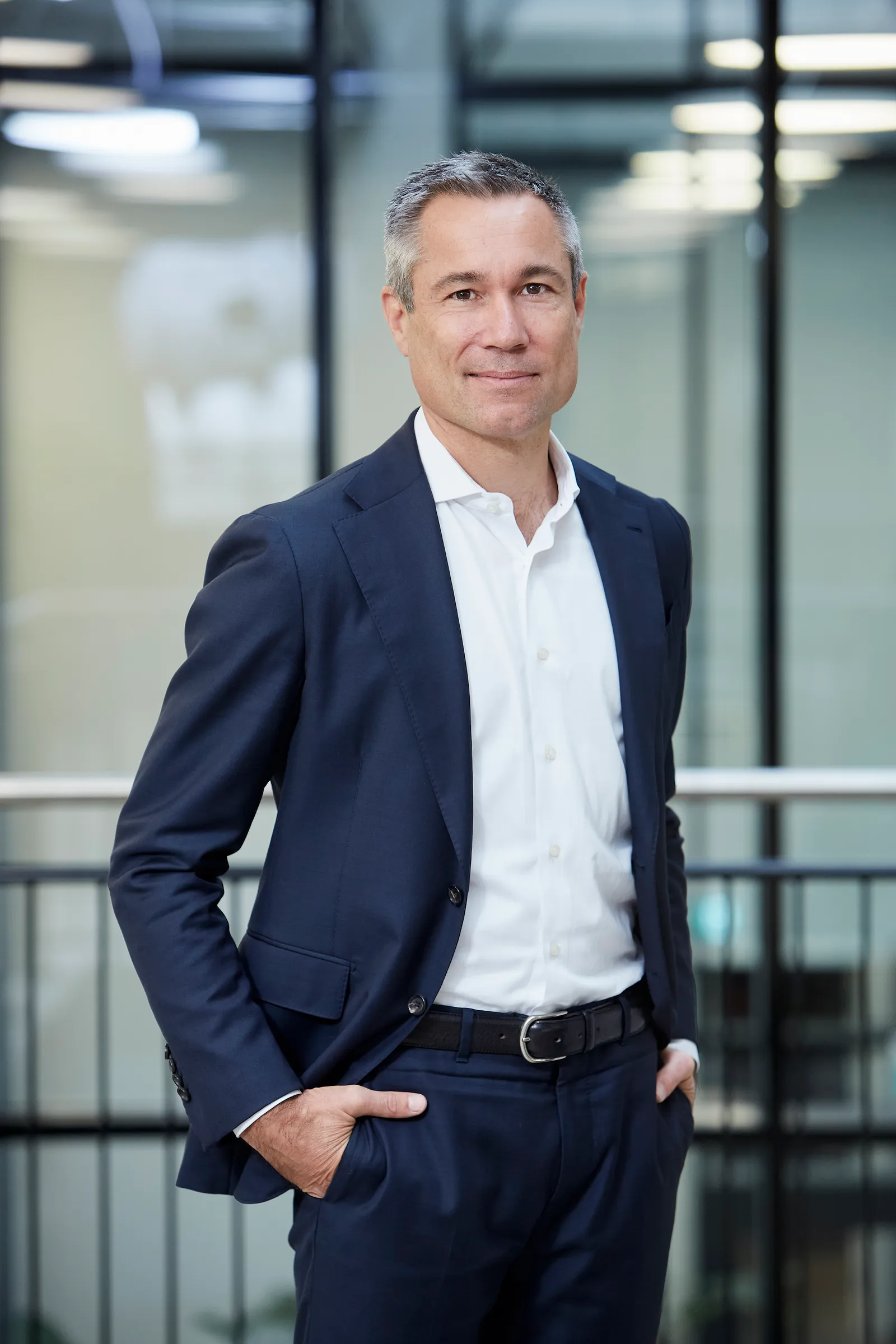 a man stands with hands in pockets of blue suit against blurred office background