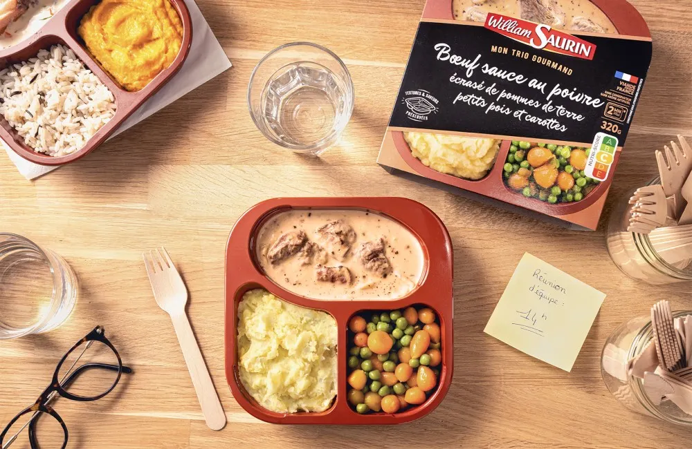 A three-compartment tray with different foods in each compartment sits on a table next to other various items.