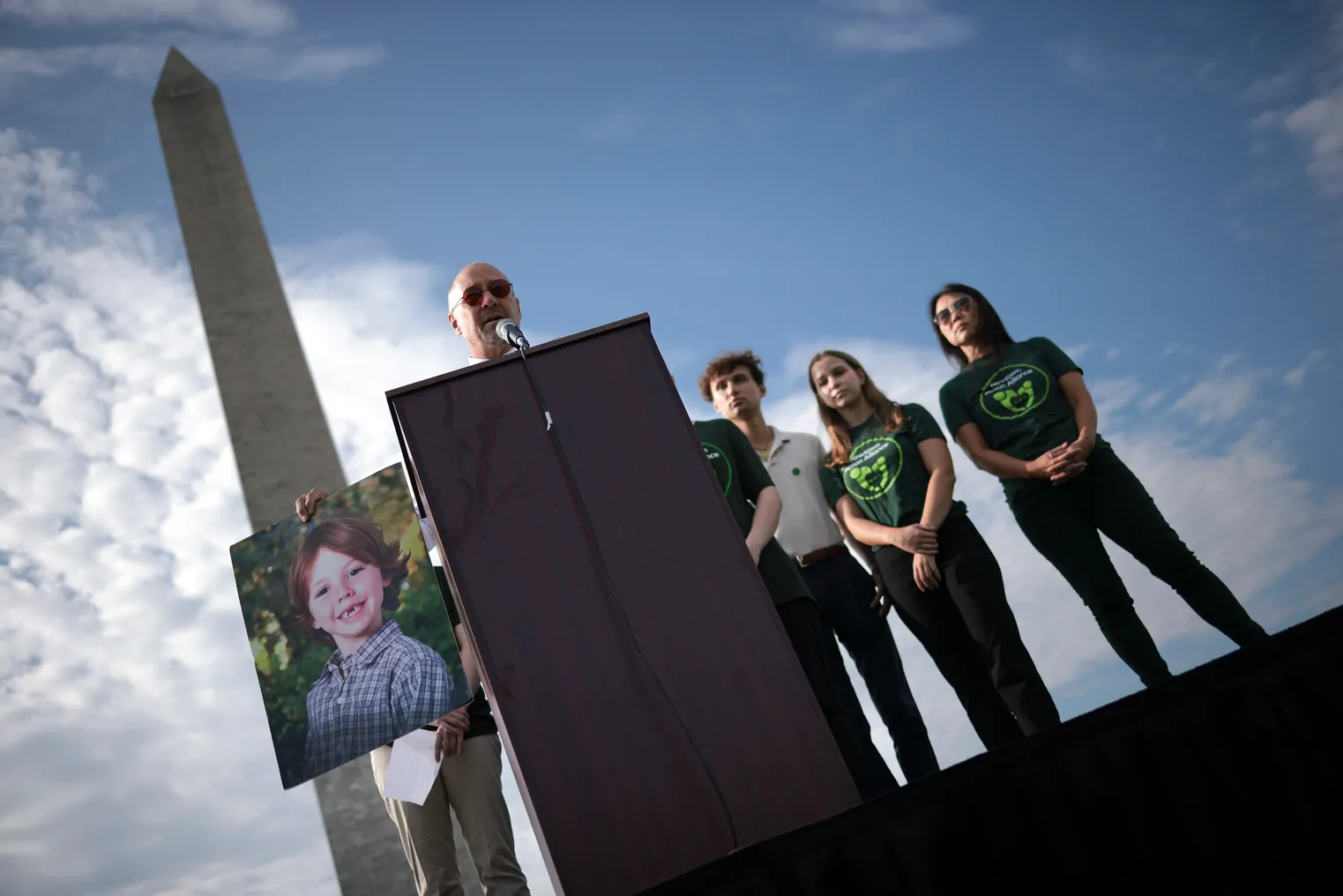 An adult stands behind a podium near the Washington Monument in Washington, D.C. To the adult's right, there is a large photo of a child. People are standing behind  the adult.