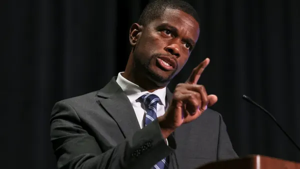 A person stands at a podium and gestures with a raised finger.
