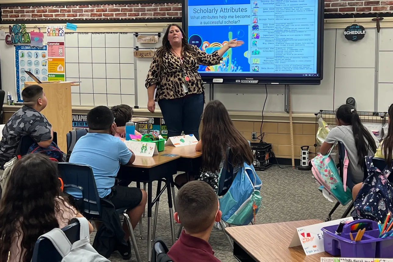 A person speaks in front of a classroom of children. Behind the teacher is a screen displaying “Scholarly Attributes.”