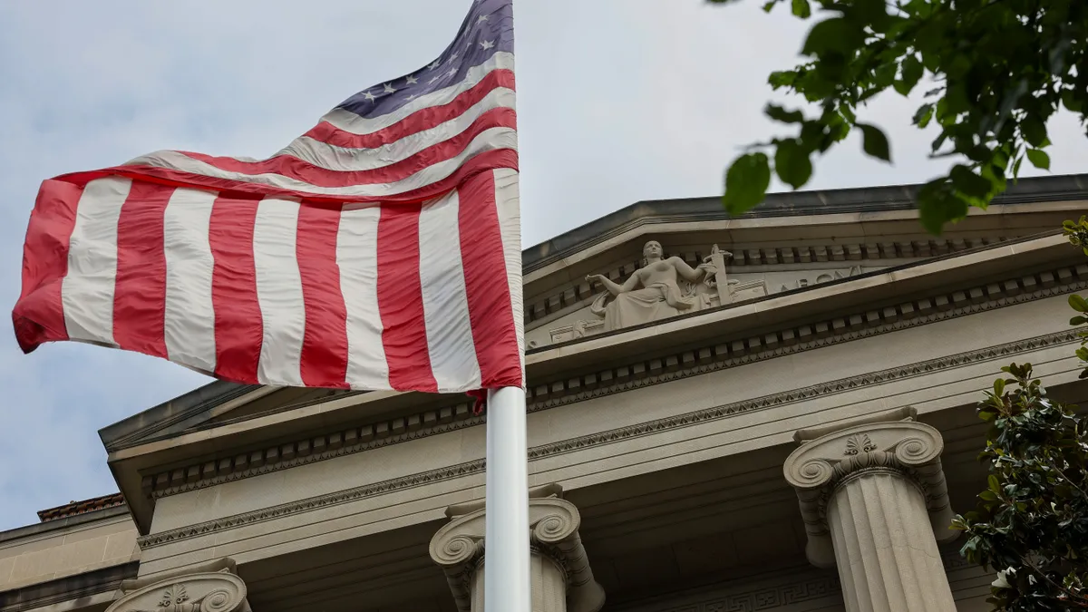 An American flag waves in front of the Department of Justice exterior. It is a neo-classical building with columns.