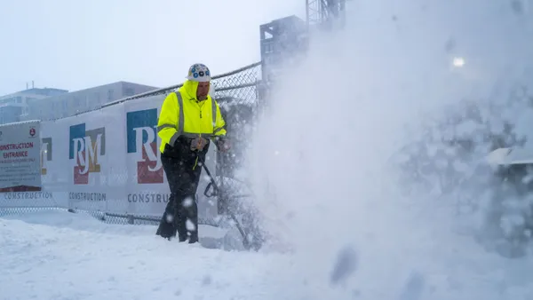 A construction worker in safety gear operates a snow blower in intense wintery conditions.