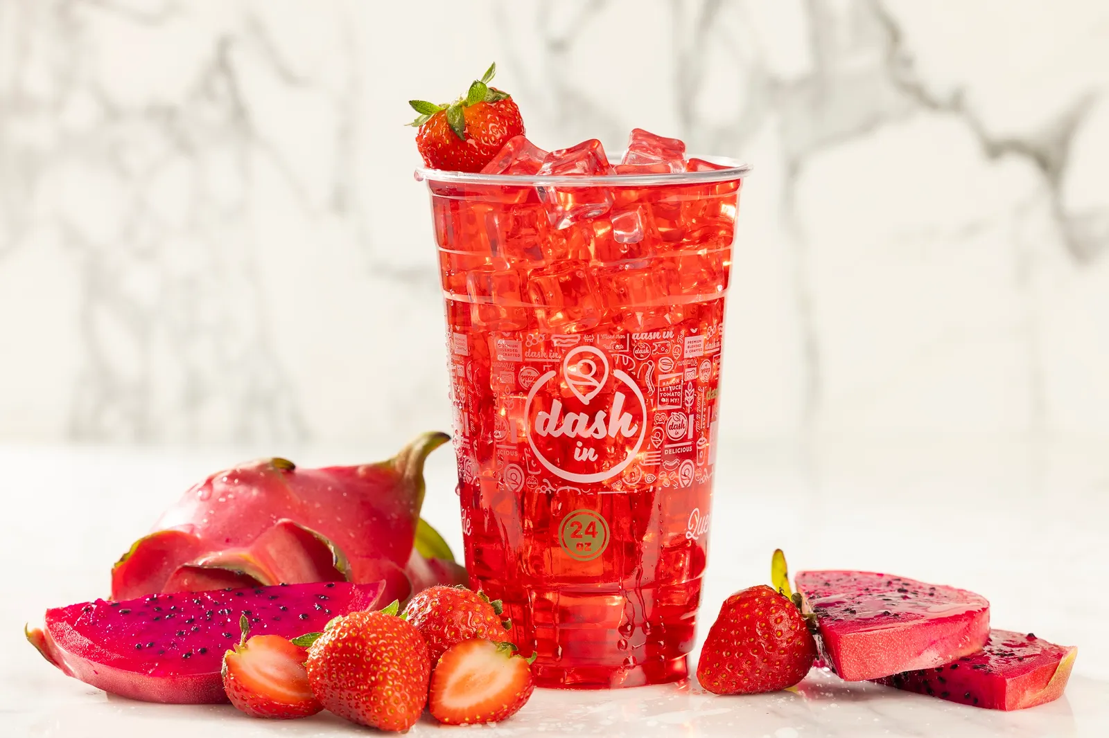 A brightly colored dink in a clear plastic cup sits on a white surface against a white and gray marble background, surrounded by fruit.
