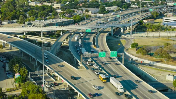 Aerial view of a major highway interchange in San Antonio, Texas, during the day. The complex multi-level freeway system features curved on-ramps and off-ramps connecting multiple interstate highways, with steady traffic flow including cars, trucks, and a school bus.