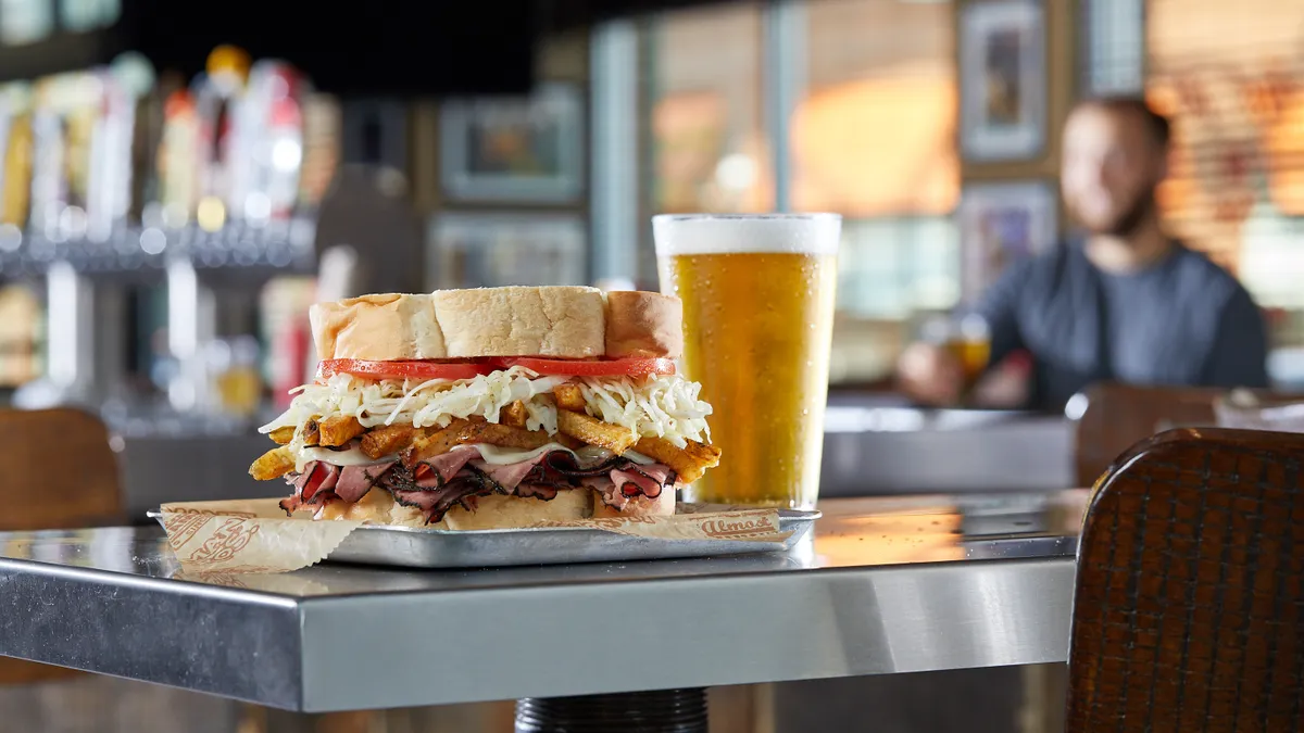 A Primanti Bros. sandwich sits on a restaurant tabletop with a diner in the background