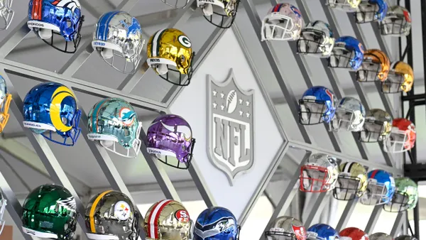 A collection of helmets for all 32 National Football League teams is on display against a wall which has the NFL shield logo at its center.