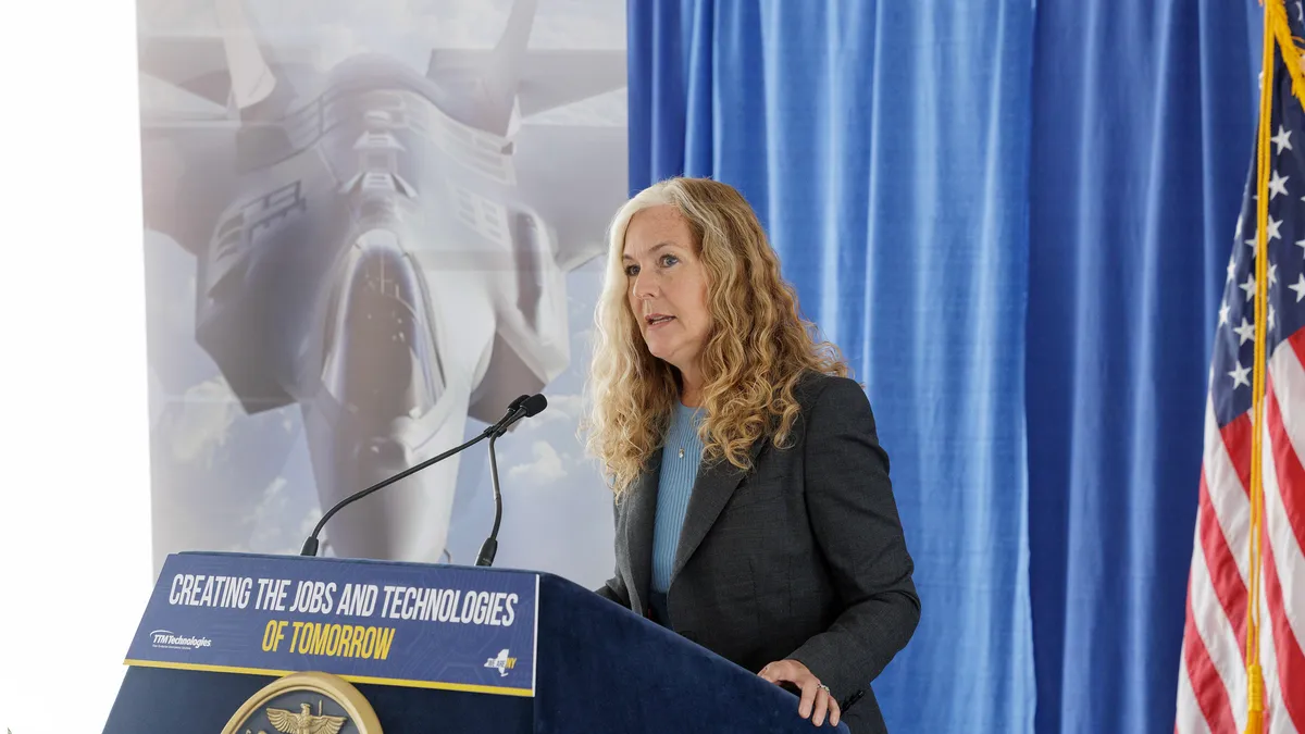 A person with wavy blonde hair standing behind a podium, with the TTM Technologies sign in the background.