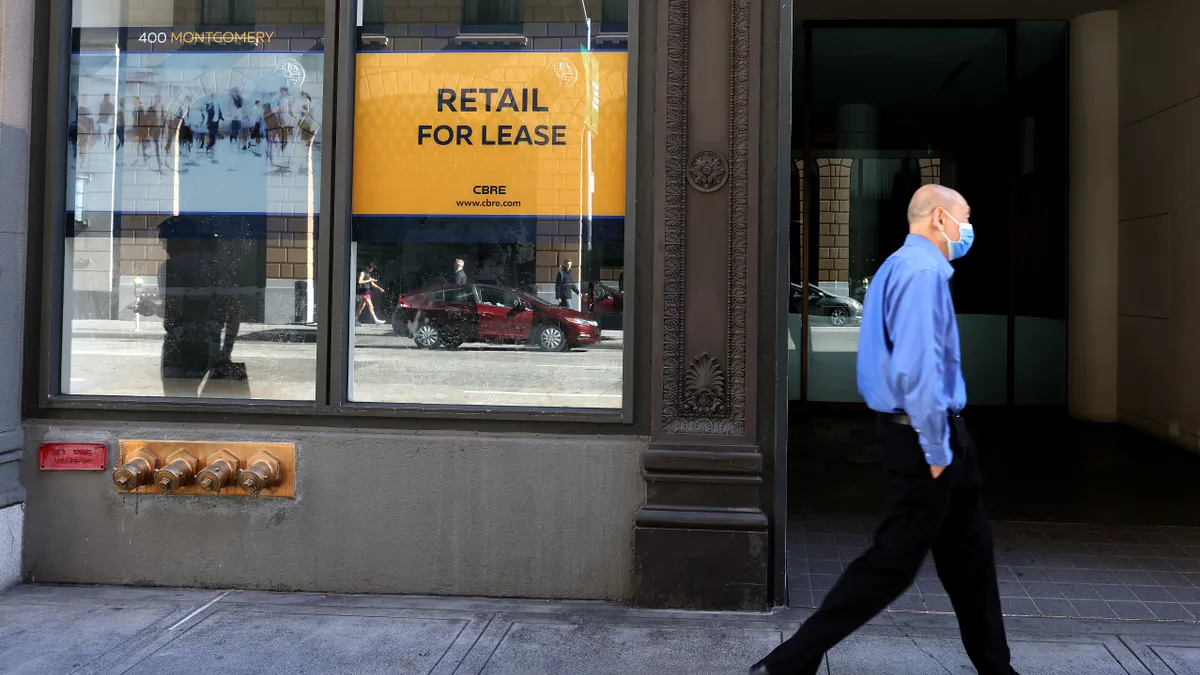 A person in a blue shirt and black pants and shoes wears a surgical face mask and walks past a window. In the window, a sign reads "Retail for Lease."