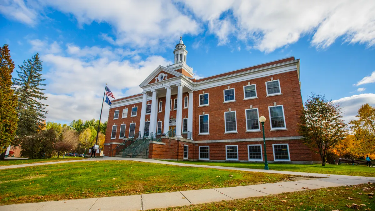 A brick academic building sits behind a lawn and in front of fall foliage.