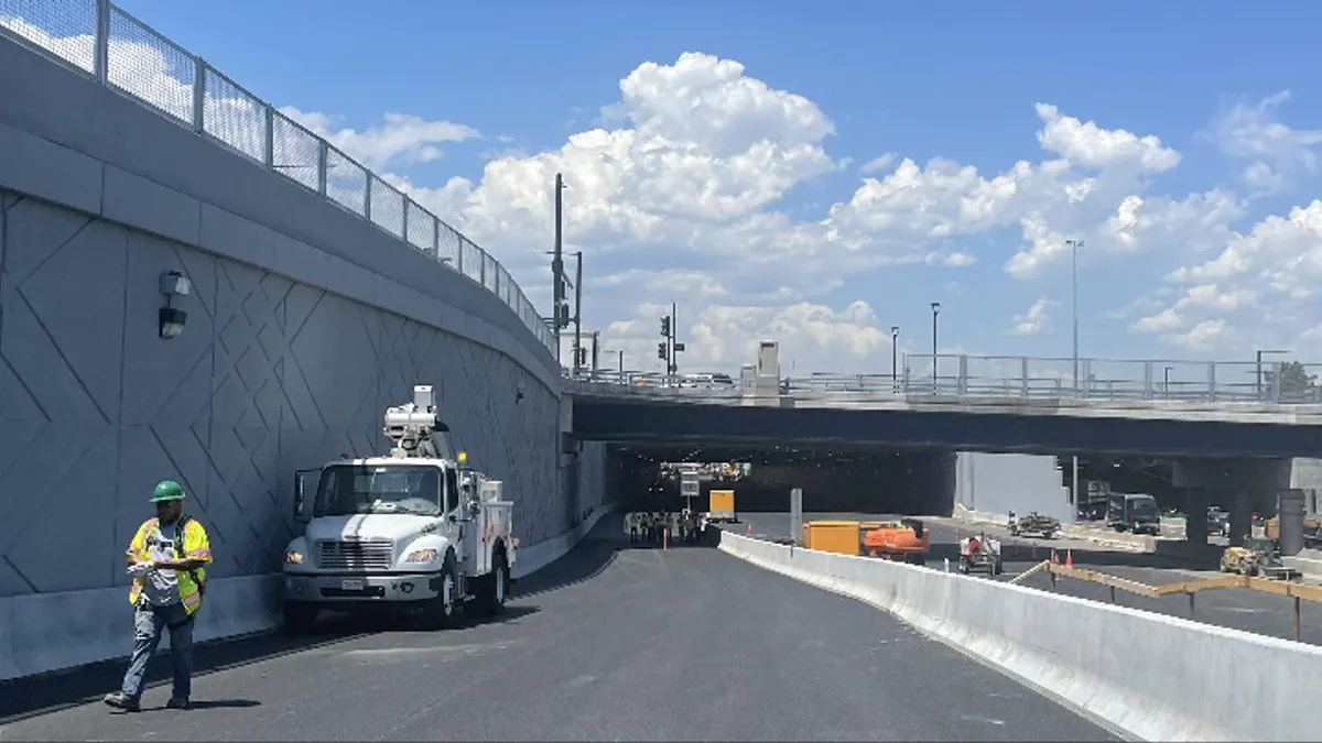 A construction worker in a reflective vest and green hard hat walks on a fresh stretch of asphalt, with an overpass in the background.