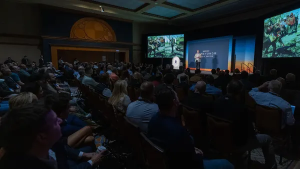 An audience sits in a darkened room and watches a person on stage give a presentation.