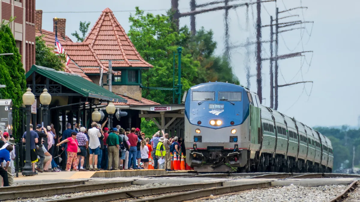 An Amtrak train at a train station with a sign reading "Manassas" and a crowd of people on the station platform.