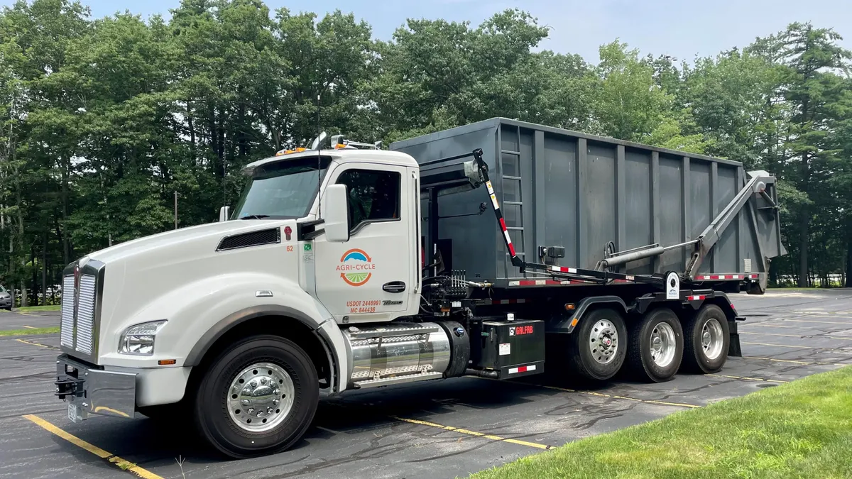 A truck with a dumpster on its back and the Agri-Cycle logo on its cab is parked in a parking lot.