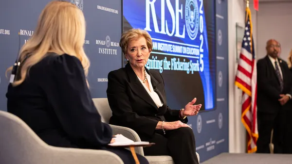 Two people are seated on a stage. One person's back is to the camera and one person is facing toward the camera. A backdrop and screen say "RISE"