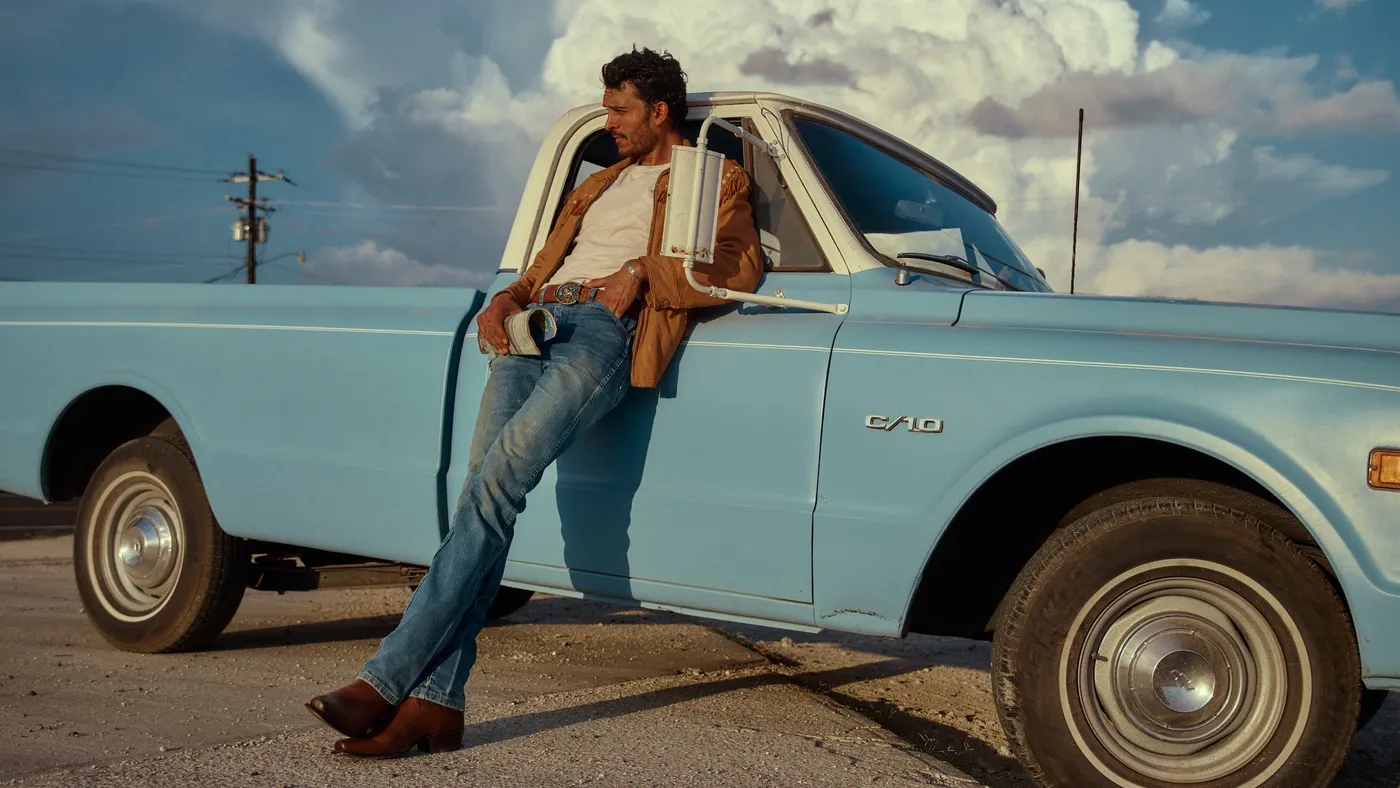 A person leaning against a blue pickup truck wearing a tan button-up shirt and blue jeans with Tecovas boots.