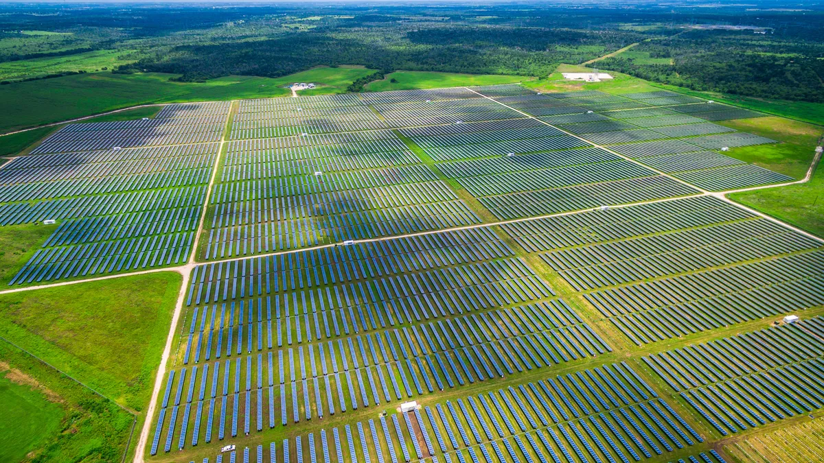 Rows of photovoltaic solar panels cross a green field with trees in the distance.