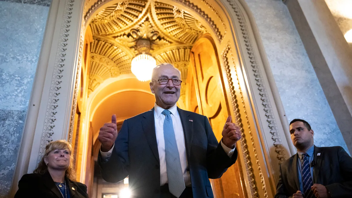 senator Chuck Schumer pauses under an arch in the United States Senate. Both of his hands are raised and giving a 'thumbs up'