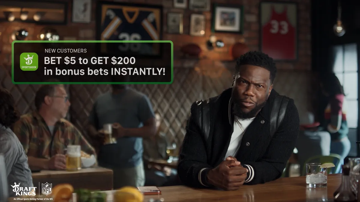 Comedian Kevin Hart sits at a bar in a black jacket in a new ad for online sportsbook DraftKings.