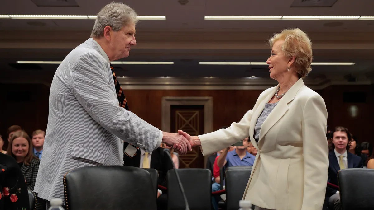 Two people are standing in a room and shaking hands. Other people are seated in rows of seats behind them.