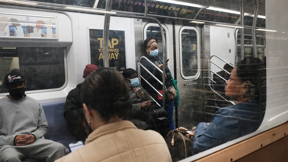 A New York subway car is seen from the outside looking through a window showing several seated and standing passengers.