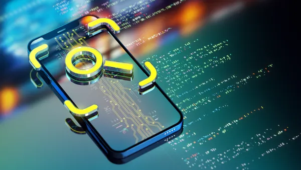A yellow loupe icon seen on top of a smart phone and data code seen on a monitor behind it.