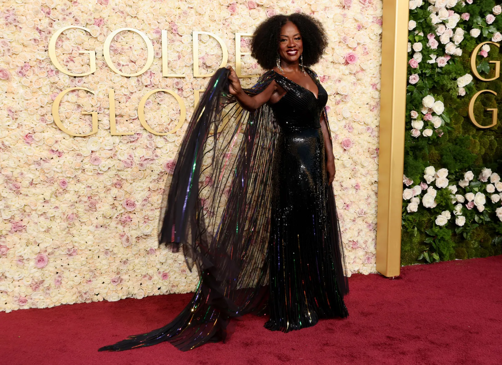 A person holds up the cape-like sleeve of a rainbow tulle dress as she stands in front of a gold sign that says Golden Globes.