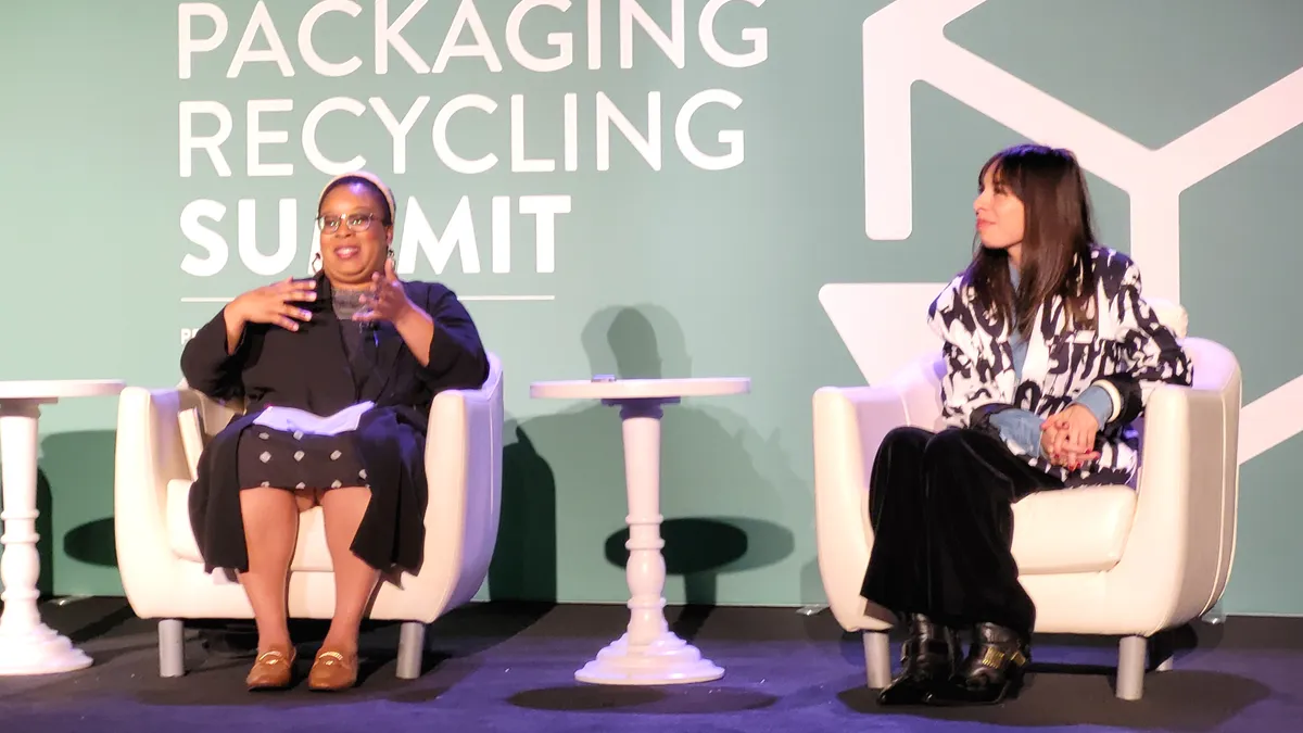 Two people sit in white chairs on a stage with a green background that says "Packaging Recycling Summit."