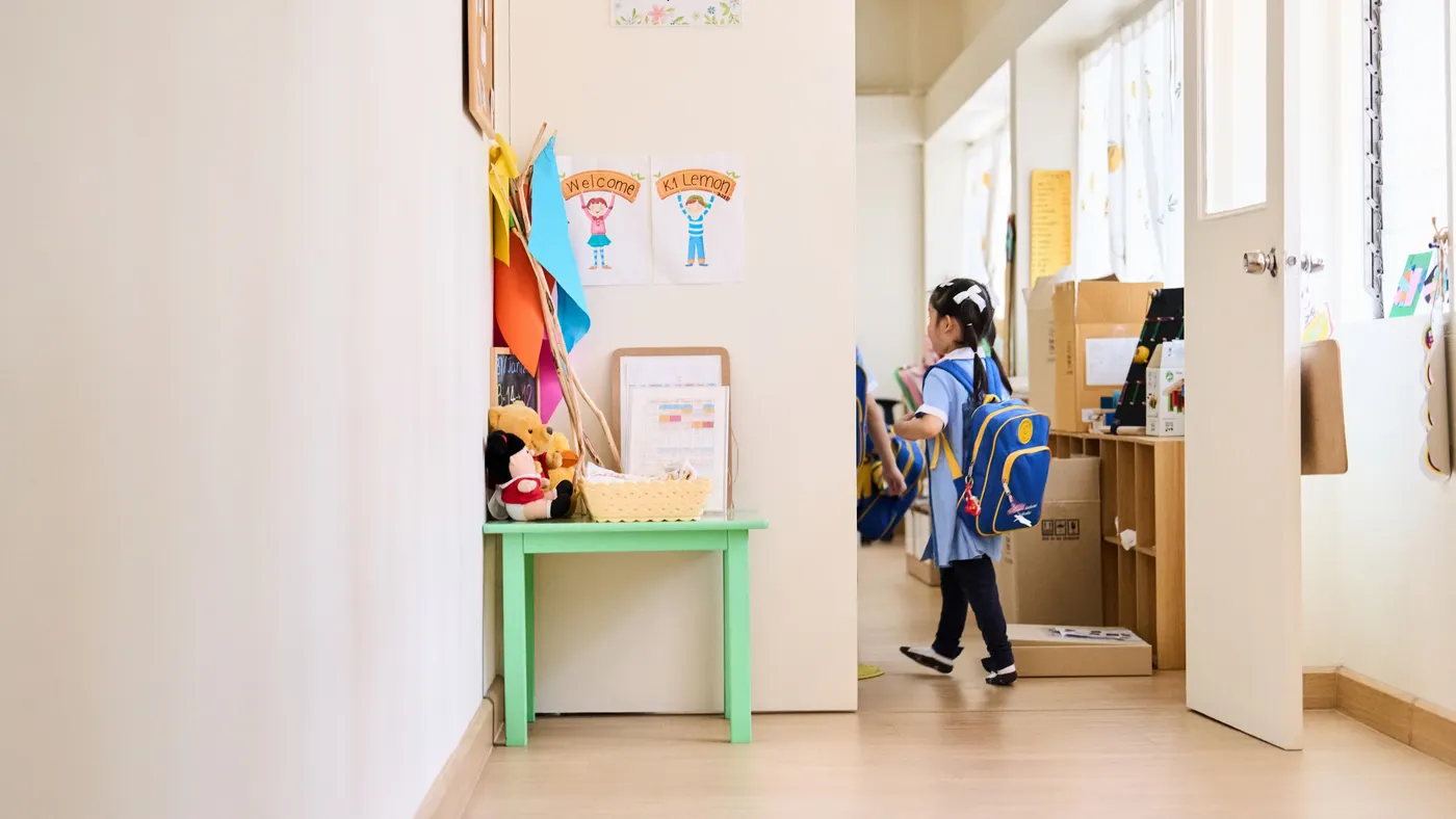 A young student wearing a backpack and holding books enters a classroom. The student is shown from behind.