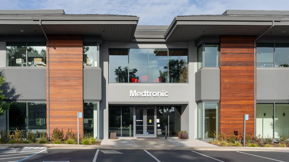 A modern two-story commercial office building with a mix of gray and wood paneling on the exterior. Large glass windows dominate both floors, and the front entrance features the "Medtronic" logo above double glass doors. The surrounding area includes a few small shrubs and empty parking spaces in front.