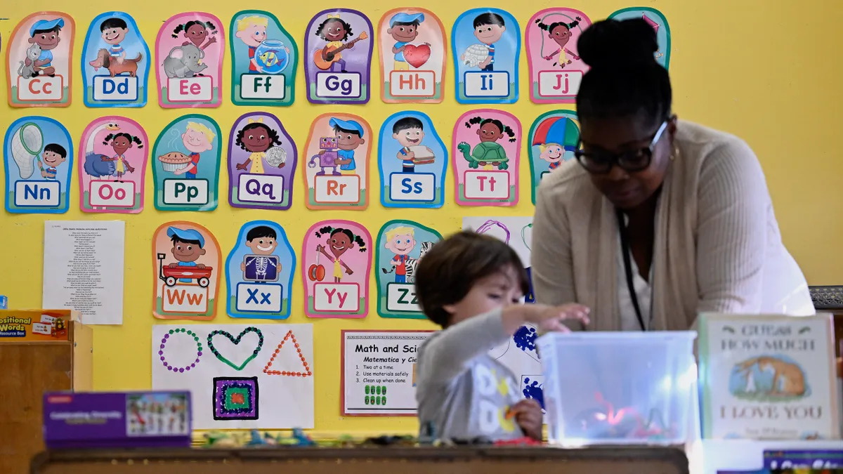 An adult is behind a desk in a room. To the person's right is a child. Both child and adult are looking at a clear container on the desk. Behind them on the wall are pictures with the alphabet.