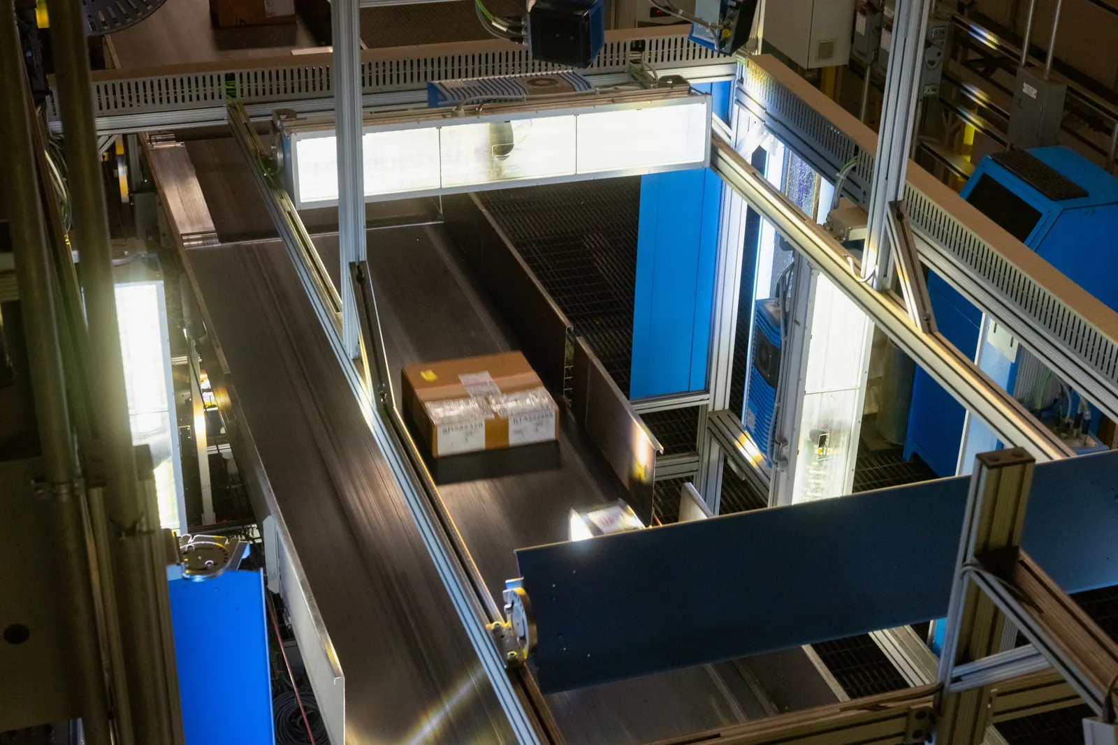 An overhead view of a cardboard package with a shipping label moving along a conveyor belt inside an industrial sorting facility. The package is passing under a large, brightly lit scanner system integrated into a network of metal tracks and machinery.