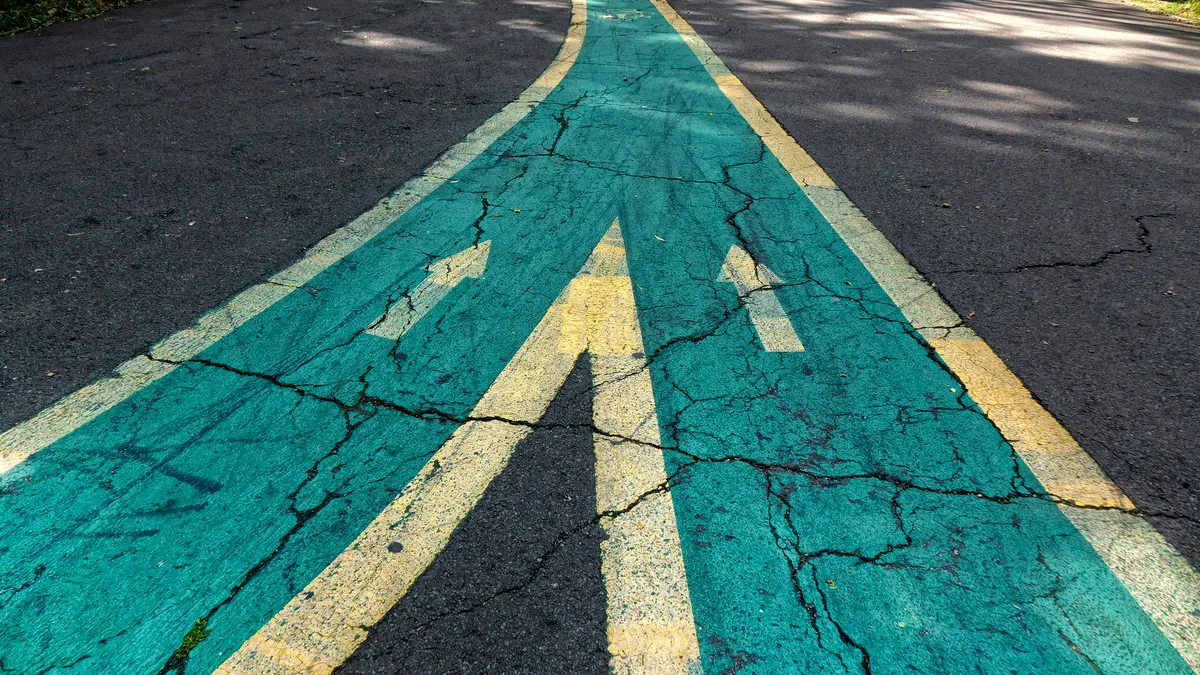 Two green roads, bordered by yellow paint, converge on an asphalt road with arrows pointing forward away from the frame.