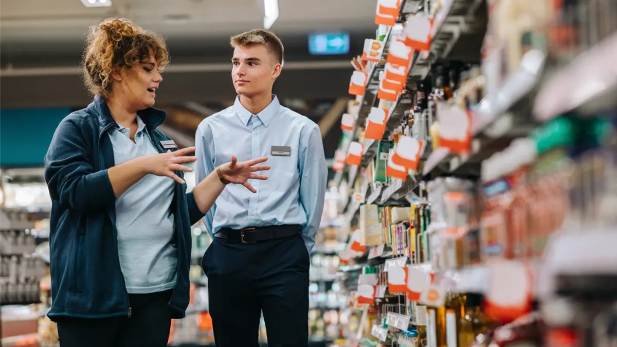 A supermarket manager is explaining something to a trainee while standing in an aisle surrounded by various products.