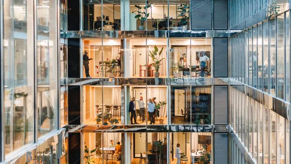 A well-lit office setting shows employees across multiple floors and rooms of the buildings through glass windows.
