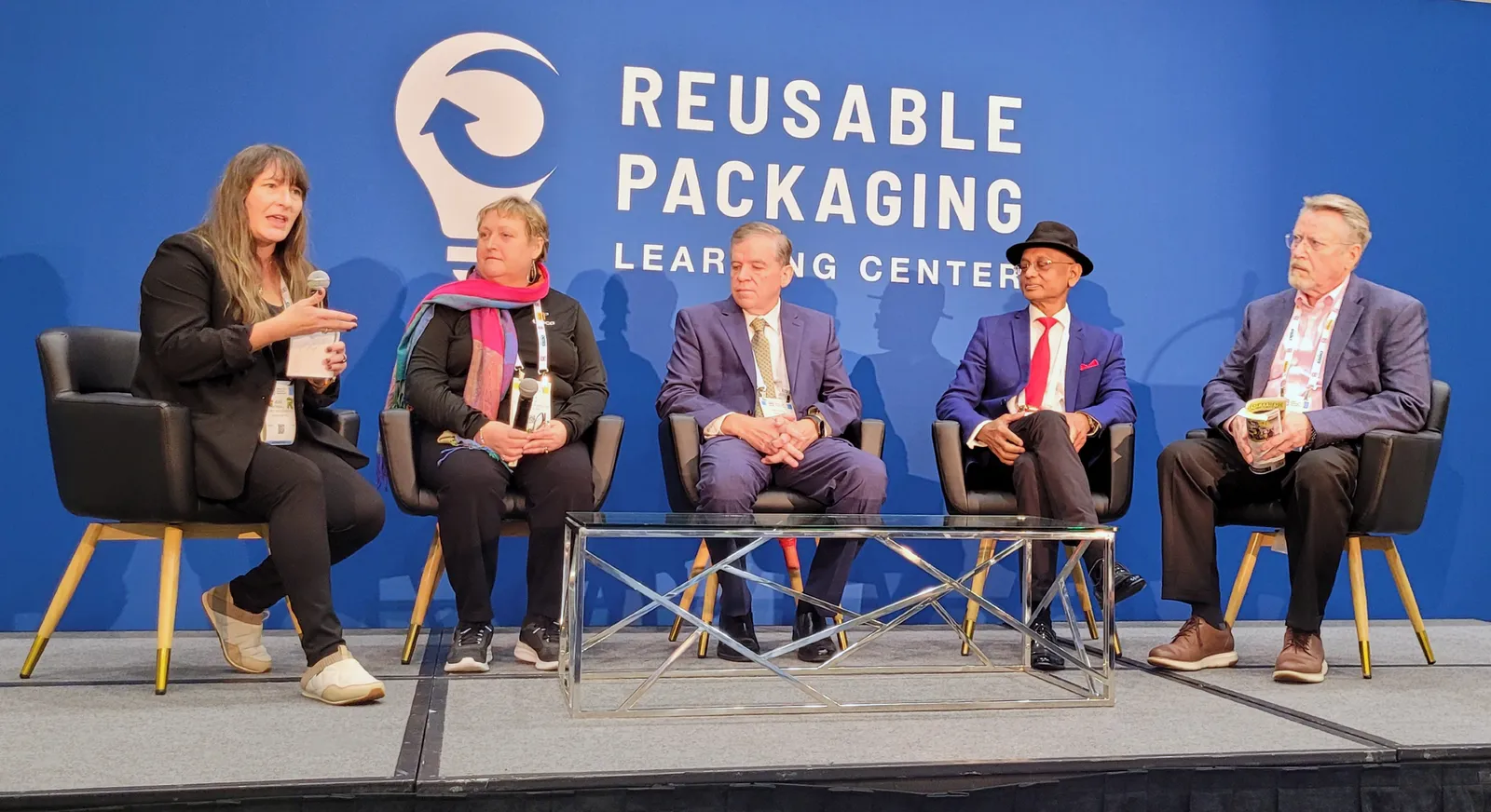 Five speakers sit in chairs on a stage at an event with a blue background that says Reusable Packaging Learning Center.