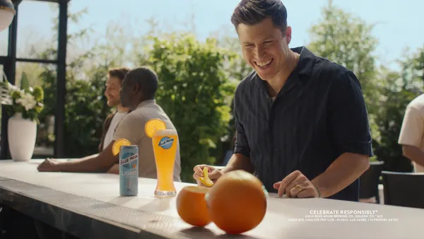 Colin Jost wears a black button-up shirt at an outdoor bar as he smiles at two oranges next to a pint of Blue Moon.