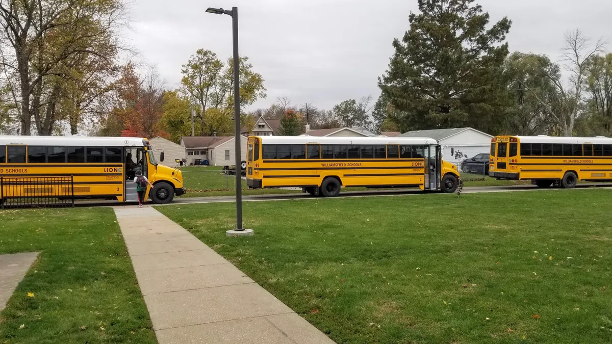 Three yellow electric school buses are lined up behind each other along a sidewalk near a grass lawn