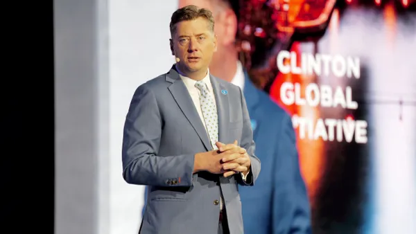 Man in gray suit with hands clasped standing in front of a blurred sign that reads "Clinton Global Initiative."