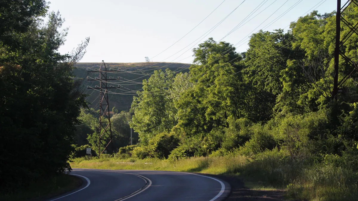 A landfill covered in grass rises behind a line of trees. In the foreground, a two-lane road bends around a curve.