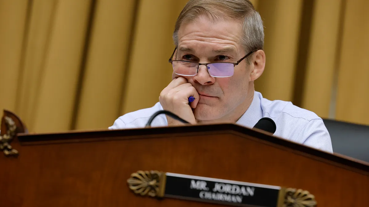 Jim Jordan sits behind a podium with his right hand around a pen and resting on his chin. Out of focus, the podium reads "Mr. Jordan, Chairman."