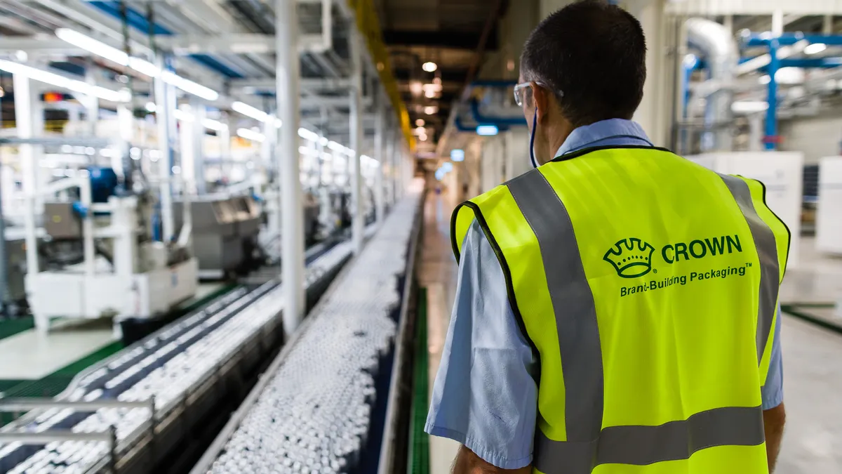 A person wearing a bright yellow vest with Crown branding on the back stands near a can manufacturing line.