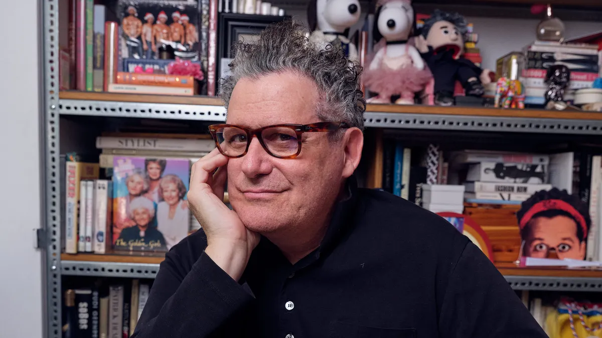 A person wearing glasses, with a hand against one cheek, looks into the camera.