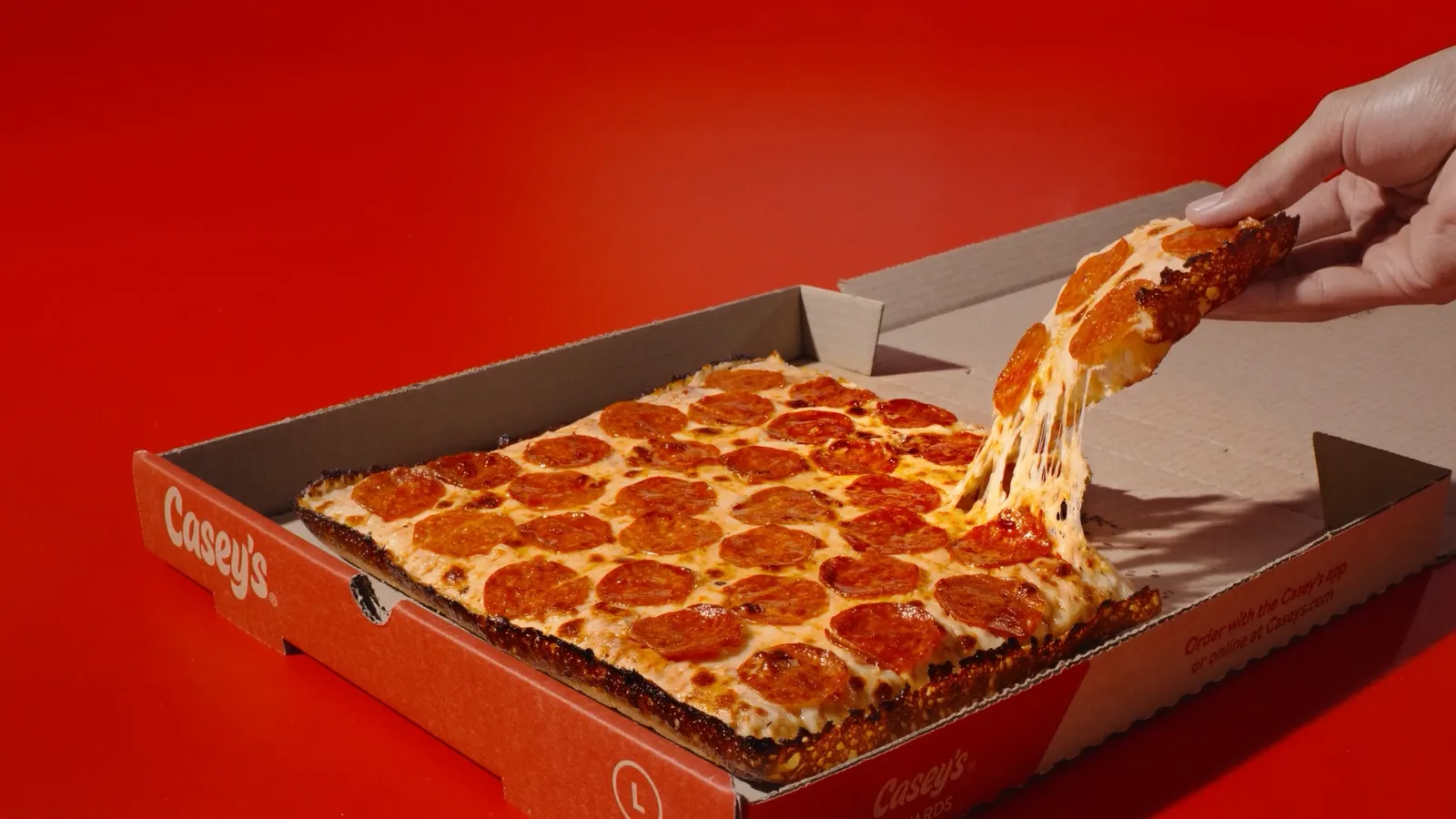 A photo of a rectangular pizza in a box against a red background. The pizza is cut into pieces and a hand is pulling one of the pieces out of the box.
