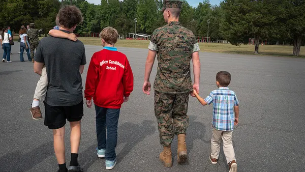 A person wearing a military uniform walks hand in hand with a child with three other children to his left