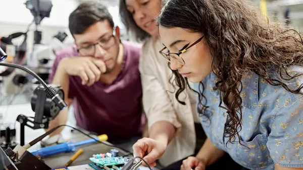 Close view of mature female educator standing between early electrical engineering students and offering guidance as they work on a project.