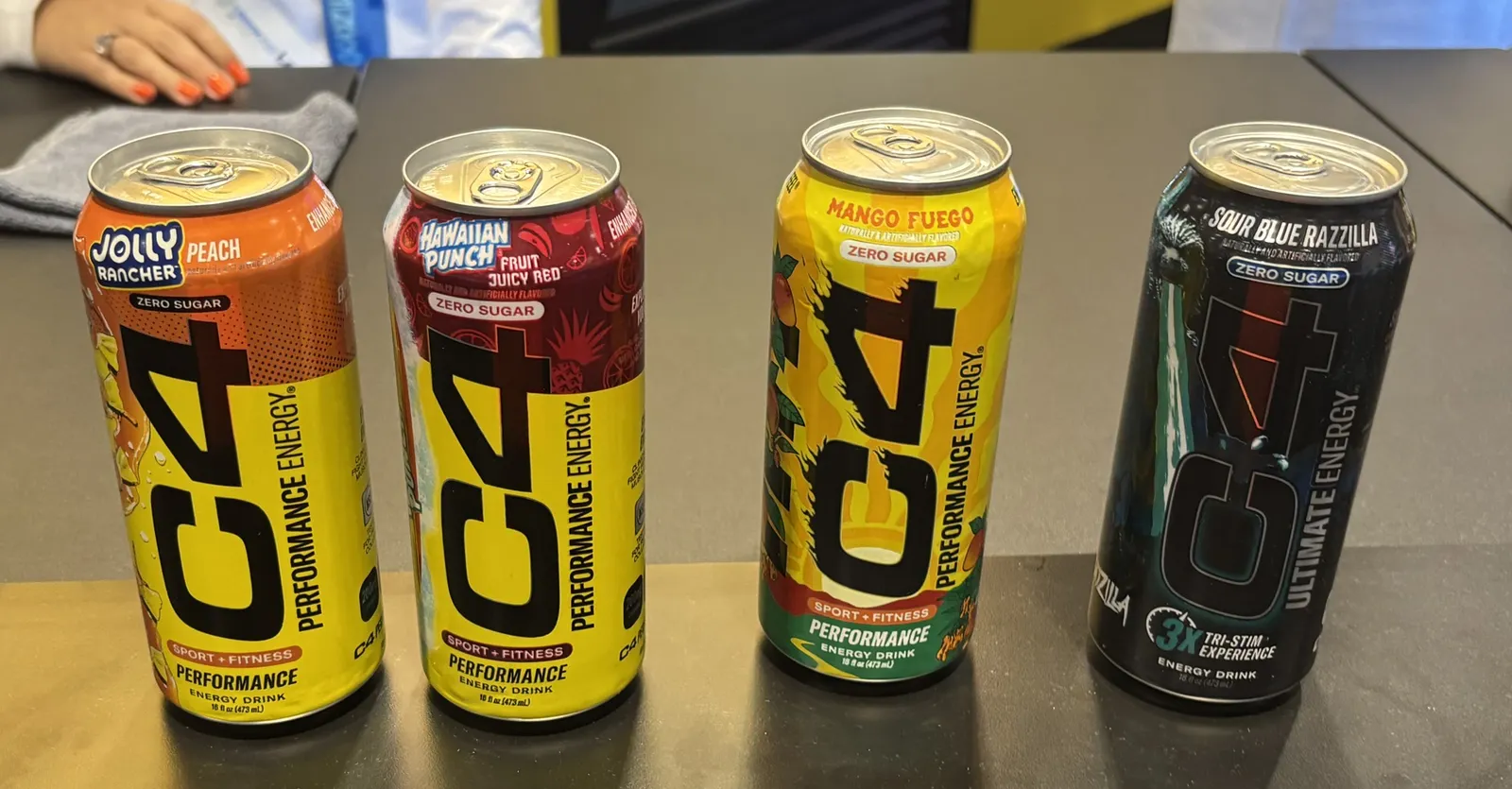 A photo of four cans against a black background. The cans say "C4" and each lists its flavor: Peach Jully Rancher, Hawaiian Punch, Mango Fuego and Sour Blue Razzilla,