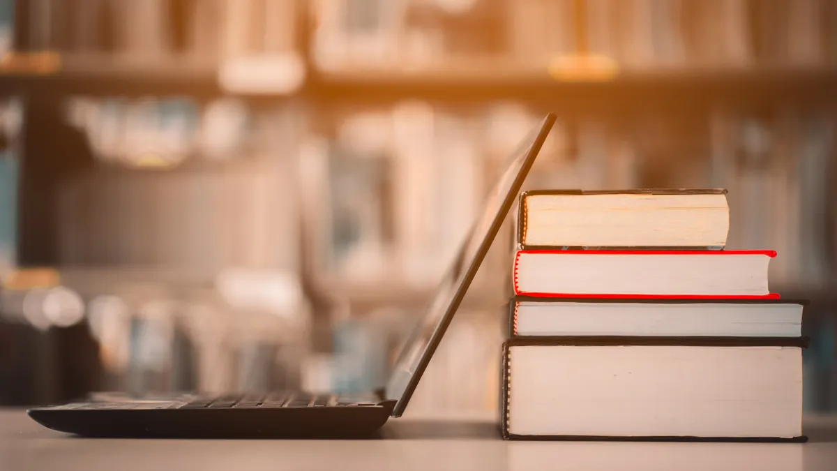 An open laptop screen rests on a stack of printed books, which are on a table. In the background seems to be a bookshelf.